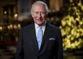 A portrait photograph of HM The King smiling as he stands in the Lady Chapel at Westminster Abbey. A Christmas tree lit with warm white lights stands in the background.