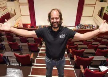 Rick Taylor with long brown hair and beard wearing black t-shirt and jeans stands at the top of tiered red velvet seats in the centre aisle of a cinema with the cinema screen behind him. He is holding his hands out and smiling.