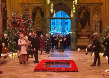 People in formal wear in Westminster Abbey, which has four Christmas trees in the background and poppies surrounding a black marble slab on the wooden floor. People are entering through the central doorway and lights can be seen outside.