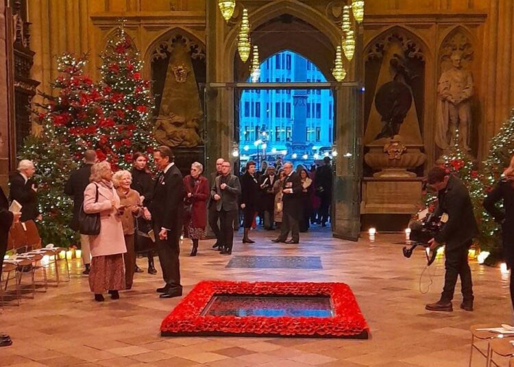People in formal wear in Westminster Abbey, which has four Christmas trees in the background and poppies surrounding a black marble slab on the wooden floor. People are entering through the central doorway and lights can be seen outside.