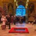 People in formal wear in Westminster Abbey, which has four Christmas trees in the background and poppies surrounding a black marble slab on the wooden floor. People are entering through the central doorway and lights can be seen outside.