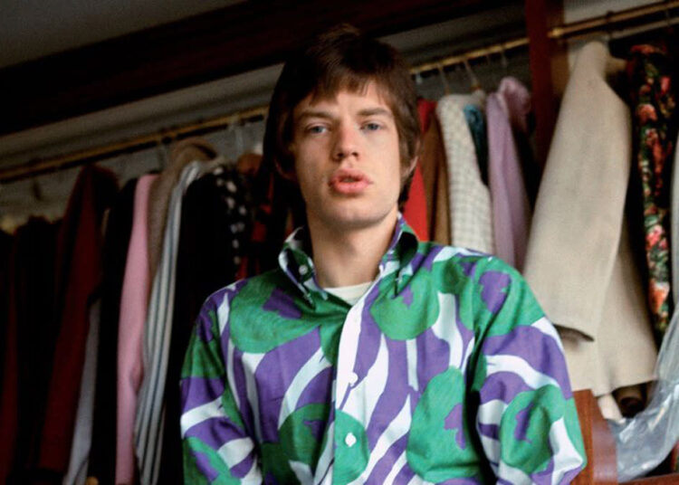 A young man with straight brown hair wears a bold, colorful shirt with green and purple patterns, standing in front of a rack of assorted clothes and jackets hanging in a wardrobe.