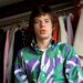 A young man with straight brown hair wears a bold, colorful shirt with green and purple patterns, standing in front of a rack of assorted clothes and jackets hanging in a wardrobe.