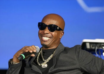 A man wearing dark sunglasses, a black jacket, and gold chains is smiling and holding a microphone, seated against a blue background with musical equipment visible behind him.