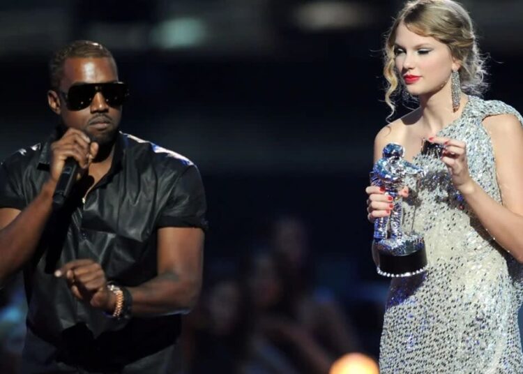 A man in sunglasses speaks into a microphone on stage while a woman in a sparkly dress holds a silver award trophy, looking surprised. The background is dimly lit with an audience visible.