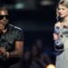 A man in sunglasses speaks into a microphone on stage while a woman in a sparkly dress holds a silver award trophy, looking surprised. The background is dimly lit with an audience visible.