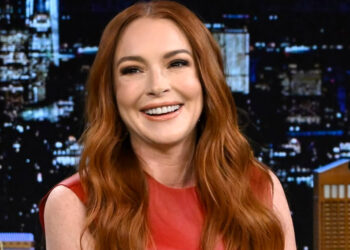 A woman with long red hair, wearing a sleeveless red dress, smiles while seated in front of a cityscape background at night.