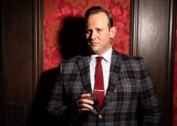 Light-skinned man in checkered suit raises eyebrow while holding lowball glass, standing in front of wall finished with polished wood and fine textile.