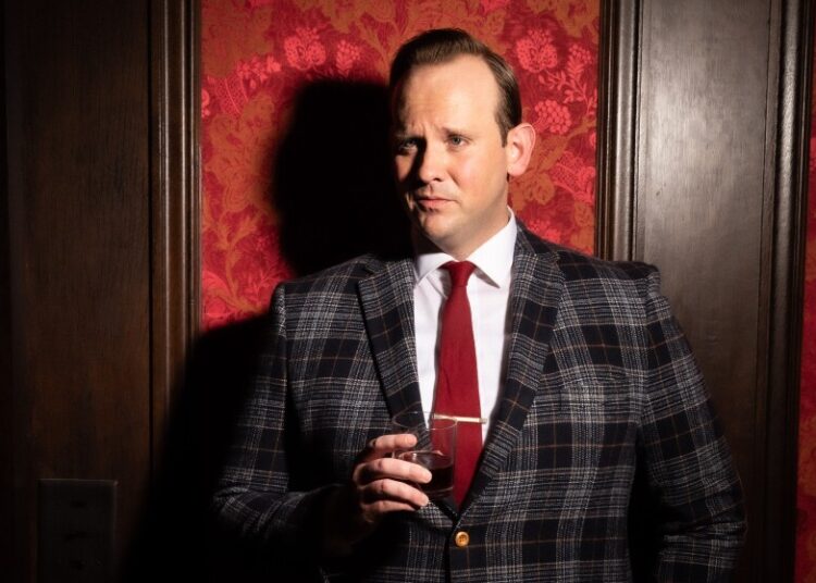 Light-skinned man in checkered suit raises eyebrow while holding lowball glass, standing in front of wall finished with polished wood and fine textile.