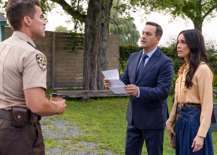 A sheriff in a tan uniform looks at a man in a blue suit and a woman an orange top and denim skirt standing next to him.