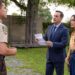 A sheriff in a tan uniform looks at a man in a blue suit and a woman an orange top and denim skirt standing next to him.