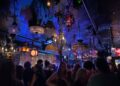 A dimly lit bar or club with ornate, eclectic hanging lamps and chandeliers, crowded with people facing a stage with colorful lights.