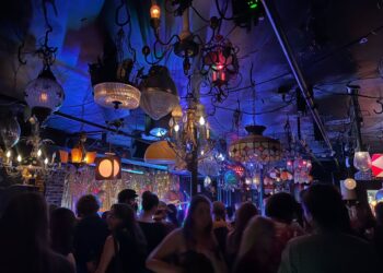 A dimly lit bar or club with ornate, eclectic hanging lamps and chandeliers, crowded with people facing a stage with colorful lights.