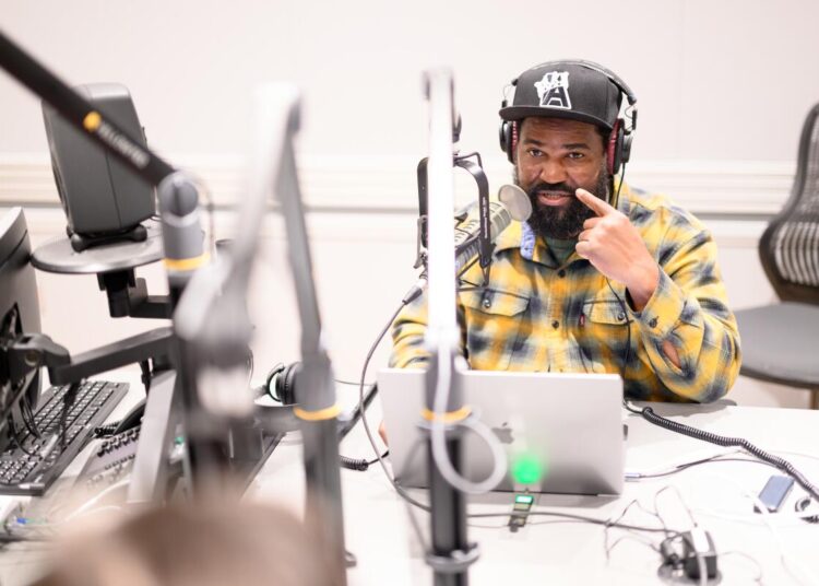 Earlonne Woods in a recording studio speaking into a mic with a laptop at his fingertips.