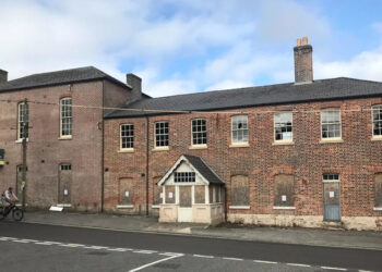 The building viewed from the other side of the road reveals it is on a hill. The ground floor windows are boarded up but the upper floor windows are exposed. There is a white timber porch with a pitched roof on the main entrance on to the pavement.