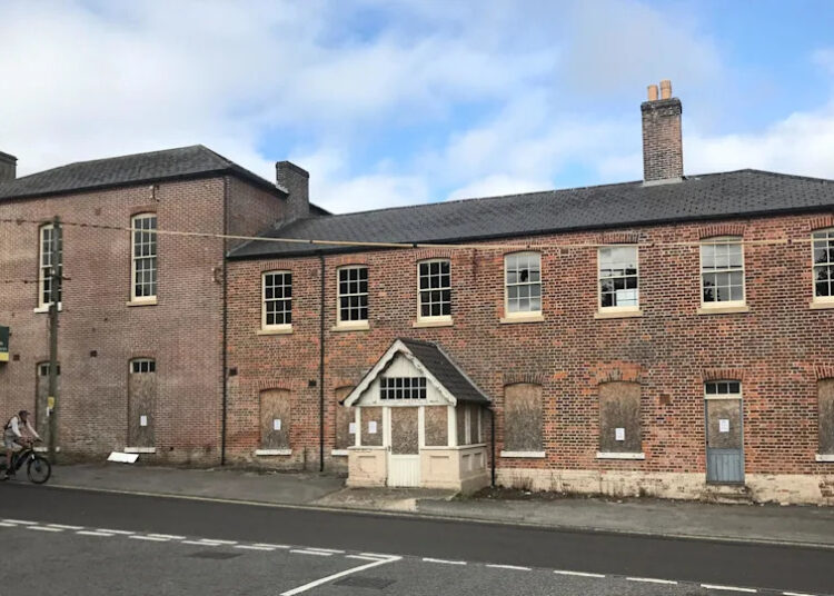 The building viewed from the other side of the road reveals it is on a hill. The ground floor windows are boarded up but the upper floor windows are exposed. There is a white timber porch with a pitched roof on the main entrance on to the pavement.