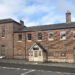 The building viewed from the other side of the road reveals it is on a hill. The ground floor windows are boarded up but the upper floor windows are exposed. There is a white timber porch with a pitched roof on the main entrance on to the pavement.