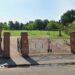 Black gates with brick pillars that open into a park which has a circular walkway and a stretch of green with various trees dotted about.