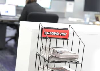 Newspapers stacked in a display stand with a "California Post" sign, in an office setting.