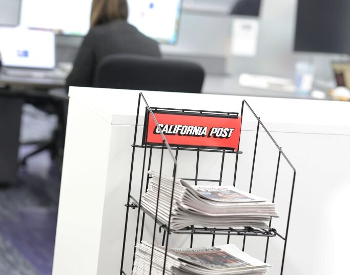 Newspapers stacked in a display stand with a "California Post" sign, in an office setting.
