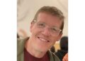 Guy Moon, in a green hoodie, burgundy shirt and clear-framed glasses, leans as he smiles.