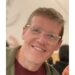 Guy Moon, in a green hoodie, burgundy shirt and clear-framed glasses, leans as he smiles.
