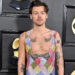 Harry Styles at an event, wearing a colorful, sleeveless checkered outfit. Black background with awards and CBS logo.