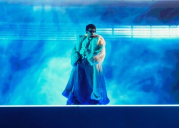 Bad Bunny performing in a white fur coat and sunglasses against a blue stage background.