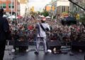 A person performs on outdoor stage. Photo taken from behind them to show large crowd on street