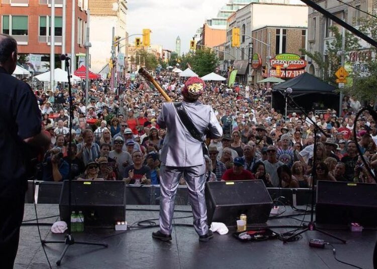 A person performs on outdoor stage. Photo taken from behind them to show large crowd on street