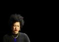 Julia Bullock in a black top with a gold and blue necklace in front of a black background