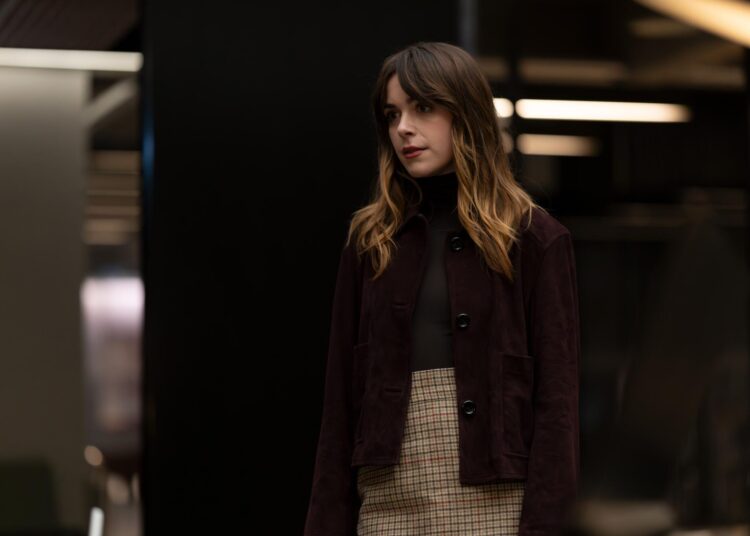A woman with long brown hair in a black turtleneck, maroon coat and plaid skirt.
