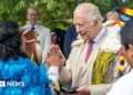 King Charles, in a suit and tie, meeting an Indigenous leader wearing a blue cloak made from feathers, at an event in Highgrove in July 2025
