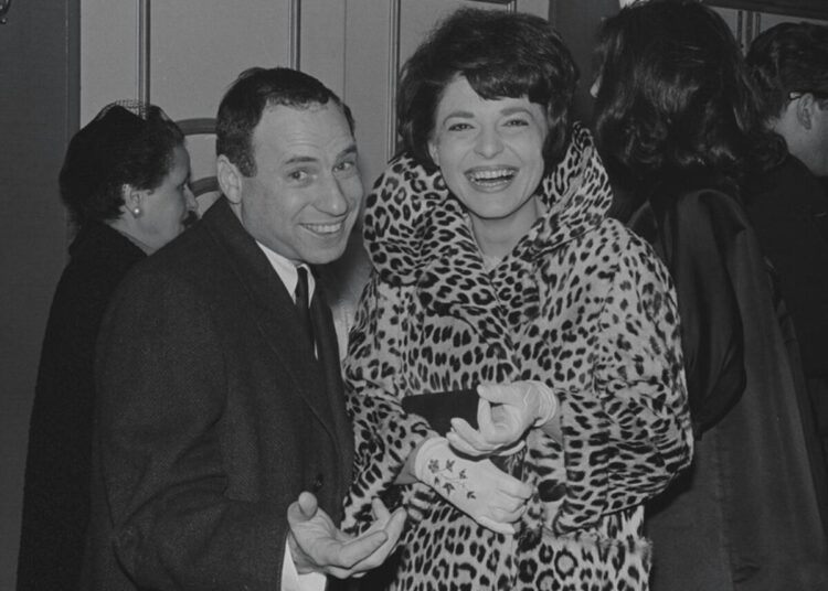 A black and white photograph of a man in a dark suit with a laughing woman in a leopard-print coat.