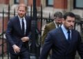 Prince Harry walks in a navy suit outside on the street behind several men