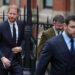 Prince Harry walks in a navy suit outside on the street behind several men