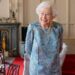 Queen Elizabeth II is standing inside a room Windsor Castle on April 28, 2022 wearing a blue dress and pearls.  She is smiling
