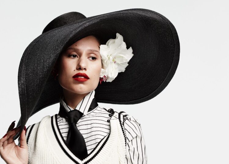 raye wearing an oversized black hat with a floral decoration