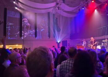 Band plays at right of photo, with its video image projected on a wall to the left, in front of a standing audience.