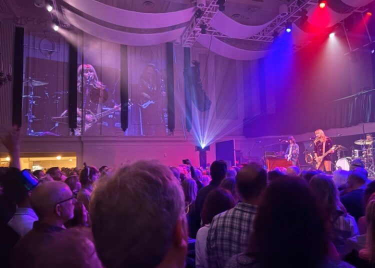 Band plays at right of photo, with its video image projected on a wall to the left, in front of a standing audience.