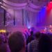 Band plays at right of photo, with its video image projected on a wall to the left, in front of a standing audience.