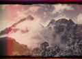 Jagged mountain peaks wrapped in mist beneath a muted sky. Red and amber light edge the image, contrasting with grays and purples in the terrain. The artist name sits at the top, while the album title, Along The Coppermine Ridge, is at the bottom.