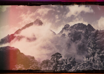Jagged mountain peaks wrapped in mist beneath a muted sky. Red and amber light edge the image, contrasting with grays and purples in the terrain. The artist name sits at the top, while the album title, Along The Coppermine Ridge, is at the bottom.