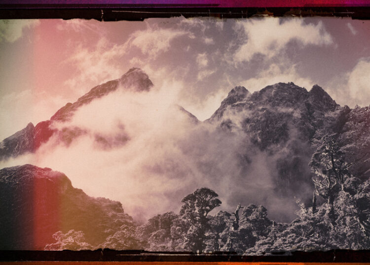 Jagged mountain peaks wrapped in mist beneath a muted sky. Red and amber light edge the image, contrasting with grays and purples in the terrain. The artist name sits at the top, while the album title, Along The Coppermine Ridge, is at the bottom.