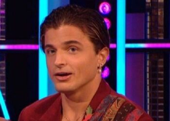 Two individuals, one dressed in a maroon coat and the other in a red coat with a scarf, are seated together on a bench, possibly on a talk show stage, smiling and engaged in conversation. The background features a multicolored lighting setup.