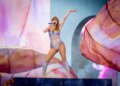 Taylor Swift performing on stage at Johan Cruijff Arena, wearing a sparkly, sequined leotard and matching boots, holding a blue microphone.