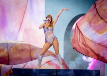 Taylor Swift performing on stage at Johan Cruijff Arena, wearing a sparkly, sequined leotard and matching boots, holding a blue microphone.