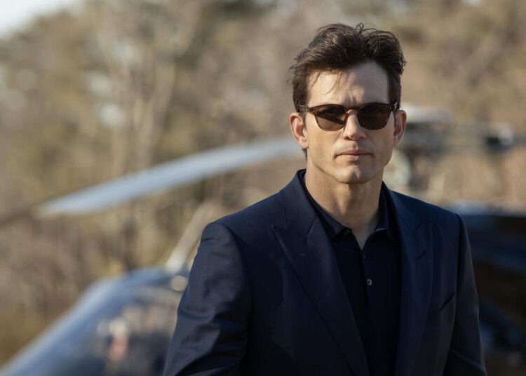 A man in a dark suit and sunglasses outside walking away from a car parked behind him.