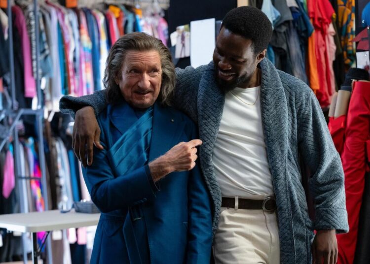 A man in a blue costume is embraced by a man in a blue robe, white T-shirt and khaki pants.