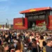 BBC An enormous crowd of people stand in front of a large stage. Massive red signs either side of the stage read 'Reading 25'. The sky is blue and largely cloudless and most of the people gathered are wearing summer clothes, with some wearing sunglasses and baseball caps.